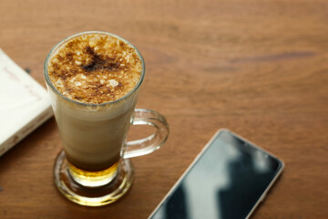 Ice coffee latte with creme brulee foam clear glass on bright wooden table. book and smartphone for decoration. Coffee time concept