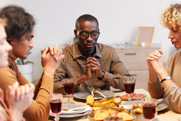 Multi-ethnic group of modern adult people praying with eyes closed while enjoying Thanksgiving dinner with friends and family