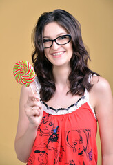 Cheerful smiling girl in glasses with a colored lollipop in her hand. Cute young brunette in a red sweater