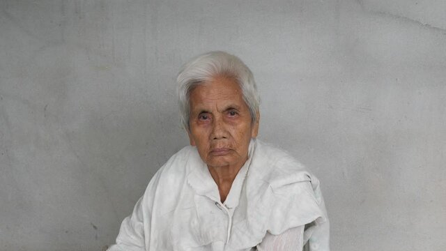 4K Asian old woman with gray hair looking to camera isolated concrete grey wall background. Rural lifestyle concept.