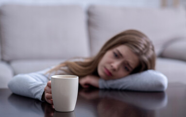 Sad young woman leaning on table and holding cup with hot drink, suffering from depression. Selective focus. Copy space