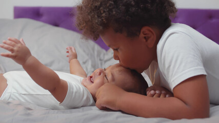 Obraz na płótnie Canvas African preschool boy kissing newborn brother lying together on bed at home