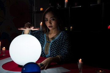 Portrait of Asian beautiful Gypsy fortune teller woman in dark room with illuminated light crystal ball