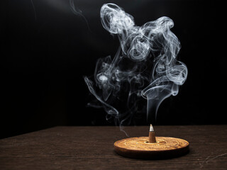Burning cone incense on wooden holder on dark surface