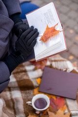Woman sits near a tree in an autumn park and holds a book and a cup with a hot drink in her hands. Girl reading a book. Fall season, leisure time and tea time concept.