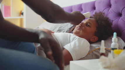 Obraz na płótnie Canvas African dad using infrared thermometer to check temperature of little sick boy lying in bed