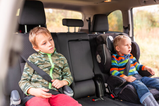 Two Little Boys Sitting On A Car Seat And A Booster Seat Buckled Up In The Car.