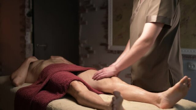 Male masseur doing sports massage of the thighs of the legs to a muscular athlete in an office with a dim light