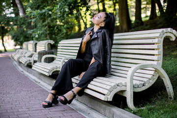 Lifestyle outdoor portrait of happy young beautiful mixed raced Asian woman, wearing black pants and leather jacket, sitting on a bench in beautiful green park. People, walk and leisure concept
