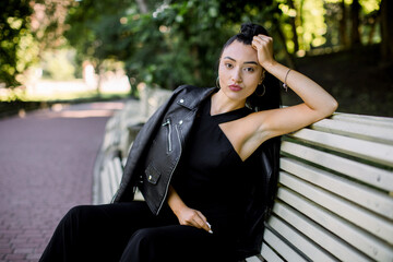 Fototapeta na wymiar Beautiful sexy Asian woman with ponytail hair style, wearing elegant black overalls and leather jacket, relaxing on bench at urban green summer park. People portraits outdoors
