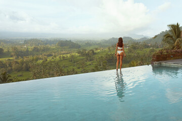 Beautiful Girl’s In Bikini In Infinity Pool Full-Length Portrait. Side View Of Young Woman With Sexy Body In White Swimsuit Posing Against Mount Agung At Tropical Resort In Bali, Indonesia.