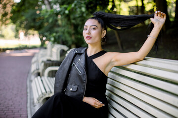 Outdoor summer lifestyle portrait of romantic young beautiful Asian woman in casual stylish black outfit, sitting on a bench in green public park, touching her ponytail, looking at camera