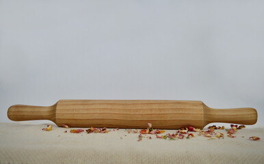 Wooden rolling pin on vanilla-colored fabric, dried rose petals scattered, white background