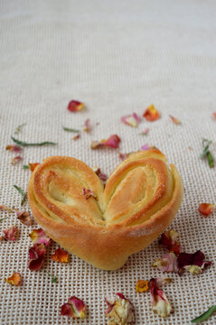 Homemade bun heart on a background of vanilla-colored fabric with scattered dried rose petals. Vertical photo