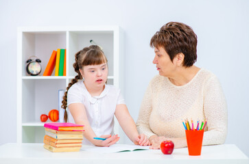 A girl with down syndrome with thick dark hair is sitting next to her teacher holding a book in her...
