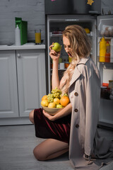 Obraz premium young woman in trench coat holding bowl with fresh fruits near opened fridge