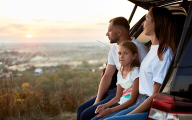 Fototapeta na wymiar Side view of a young family sitting in the car trunk enjoying the sunset outside the city, girl with a straw in her mouth is looking away, copy space