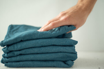 A stack of t-shirts of the same color on a light background. A man's hand picks up the top t-shirt from the stack. The minimalistic concept. Space for text.