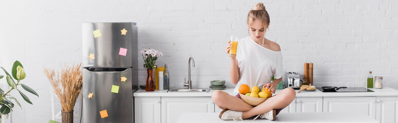 horizontal concept of young blonde woman sitting on kitchen table with fresh fruits and orange juice
