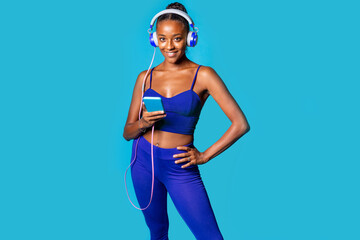 Sportive young black woman holding smartphone smiling camera on blue background - Isolated diverse african female preparing for training