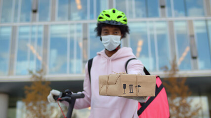 Portrait of afro delivery guy in safety mask giving you parcel standing outdoors
