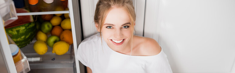 horizontal image of young woman smiling at camera near fridge with fresh fruits