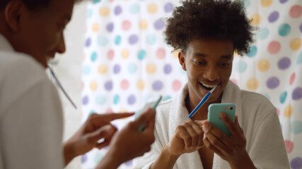 Afro man in bathrobe brushing teeth and using mobile phone in bathroom