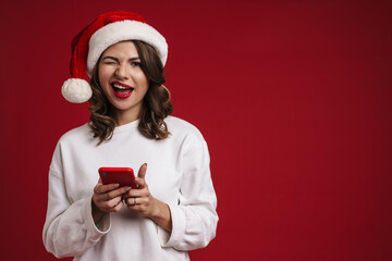 Nice happy girl in Santa Claus hat winking and using mobile phone
