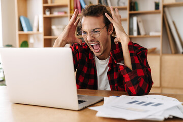 Handsome irritated guy in eyeglasses screaming while working with laptop