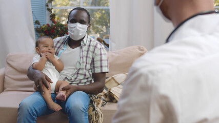 African-american dad in mask holding cute baby and listening to doctor during medical checkup at...