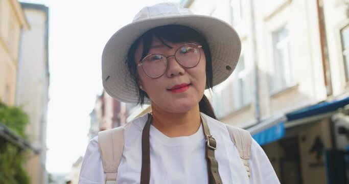 Portrait of happy middle-aged Asian female tourist in glasses and hat standing in town outdoor and smiling. Beautiful joyful woman in city on street on sunny day. Summer tourism. Close up concept