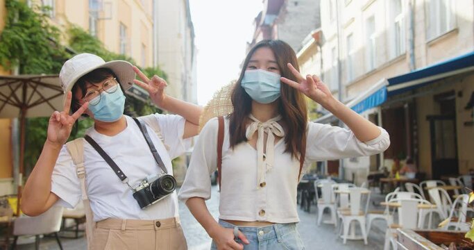 Portrait of happy mother and daughter standing on street in medical masks and posing to camera in good mood. Beautiful Asian woman with photo camera and young female tourist in city. Travel concept