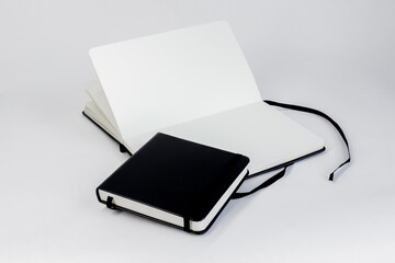 Two black notebooks. Small closed square notepad lie on large open unfold notebook with blank white pages and dark ribbon bookmark