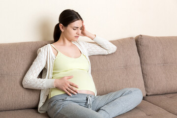 Young pregnant woman with headache sitting on sofa. Pregnancy expectation concept