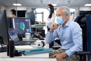 Thoughtful Senior businessman wearing face mask sitting on his desk at modern office