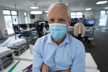 Fototapeta na wymiar Portrait of senior businessman wearing face mask with arms crossed at modern office