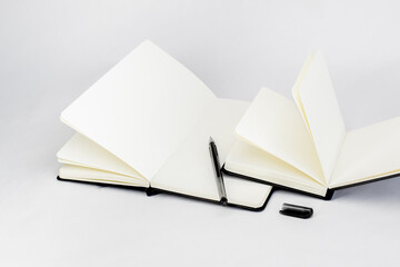 Black open small notepad and gel pen lie in unfold large notebook with blank light beige pages. White background.