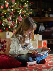 Happy young girl reading a story book in a cozy dark living room on Christmas eve. Celebrating Xmas at home. Christmas tree on the background.