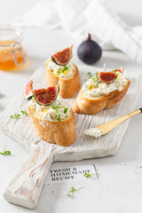 Fig, cream cheese and honey sandwiches. Canape or crostini with toasted baguette, cream cheese, figs and microgreen on a wooden board on white background. Italian recipe menu. Side view, close up