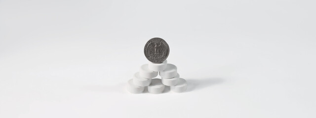 The coin stands on a mountain of pills, isolated on a white background, copy space. Pyramid of pills and a quarter dollar, banner. Medicine and health care budget, drug prices, treatment costs.