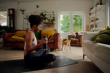 Smiling young african woman holding bottle and using smartphone during workout while sitting on fitness mat at home