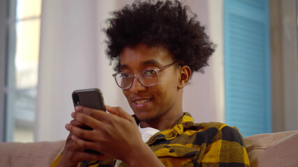 Handsome young afro man sitting on couch reading messages on mobile phone.
