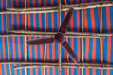 Rotating fan with slightly blurred blades on the background of the fabric ceiling in red and blue stripes and rough wooden sticks tied with ropes