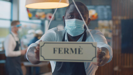 African waiter in safety mask turning ferme sign on french cafe glass door
