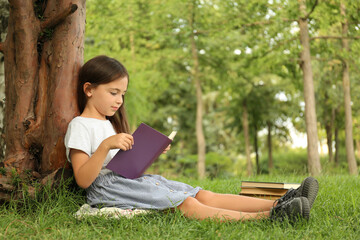 Cute little girl reading book on green grass near tree in park