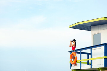 Female lifeguard with binocular on watch tower against blue sky - Powered by Adobe