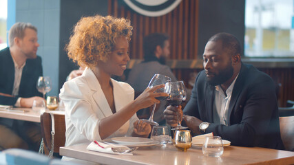 Young loving african couple sitting in modern restaurant smiling and clinking glasses with wine