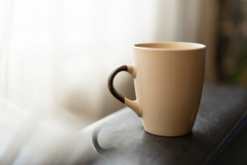 Silhouette of beige ceramic coffee mug placed on dark table near window. Blurred background with reflection of a mug and free space for copy text. Selective focus, natural day light. Low key image.