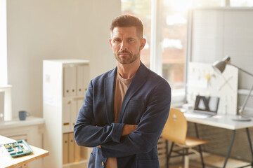 Waist up portrait of handsome mature businessman looking at camera while standing with arms crossed in office lit by sunlight, copy space