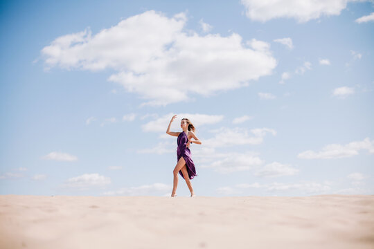 A young, slender girl in a beige dress with purple cloth in her hands posing in the desert in the wind